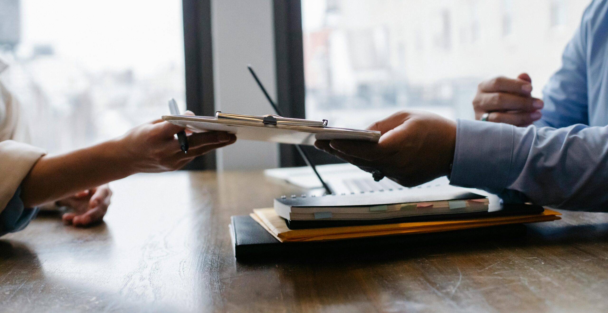 Two individuals engage in a professional exchange, one passing a clipboard to another across a wooden table strewn with notebooks, pens, and work paraphernalia.