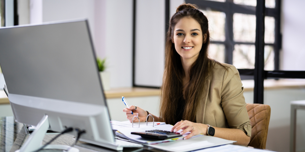A smiling young woman at a workstation with multiple devices, including a computer, calculator, and documents.