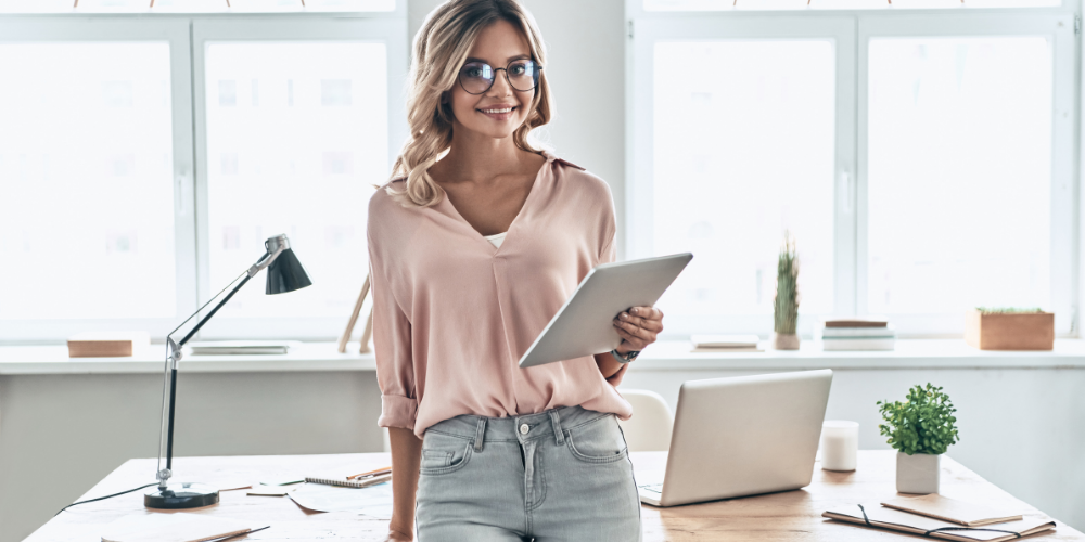A professional woman in an office standing by a desk with a tablet in hand, smiling, with a laptop and lamp in the background, symbolizing a comfortable work environment.