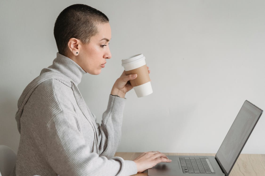 A side profile of a woman in a high-neck sweater holding a takeaway coffee cup, working on a laptop.