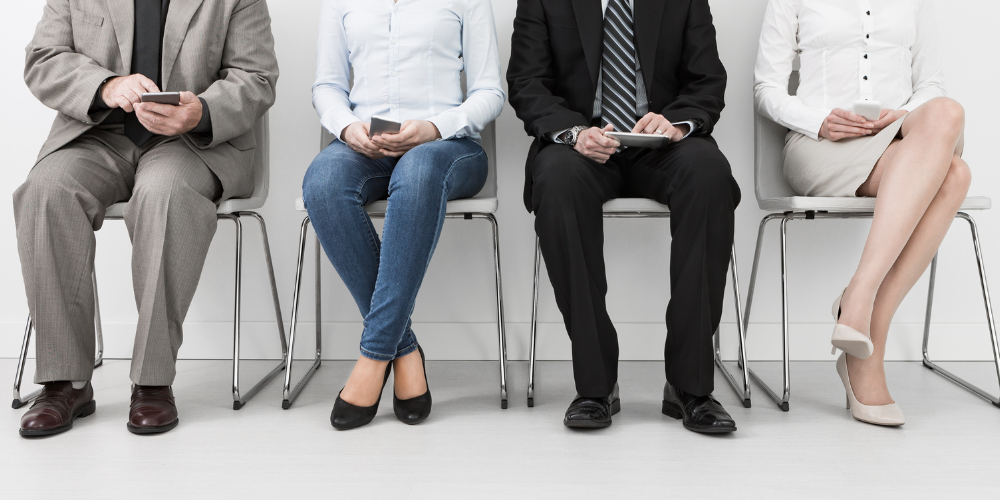 Four people seated in a waiting area, appearing to be candidates awaiting job interviews, with a focus on their lower halves and showing varied postures and attire.