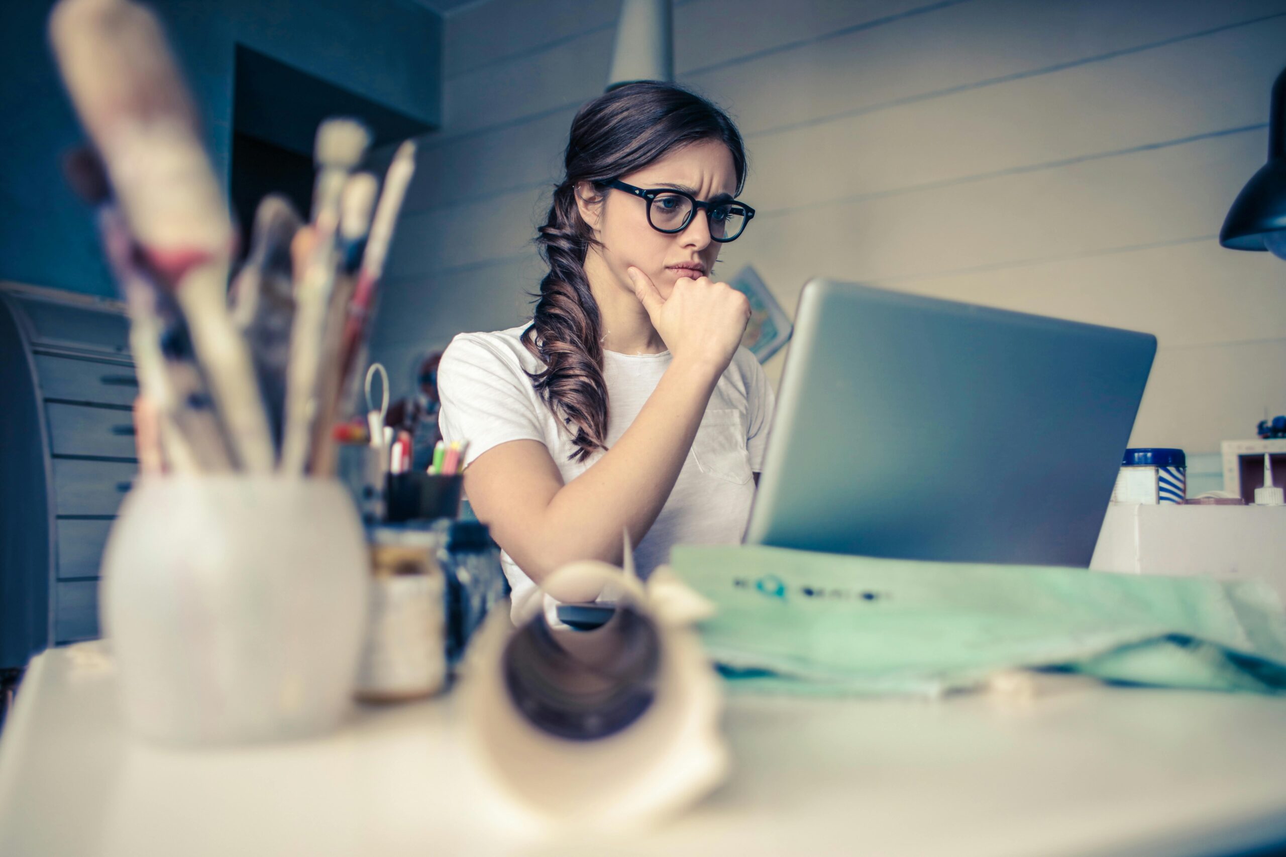 A focused woman wearing glasses looks at a laptop, contemplating."