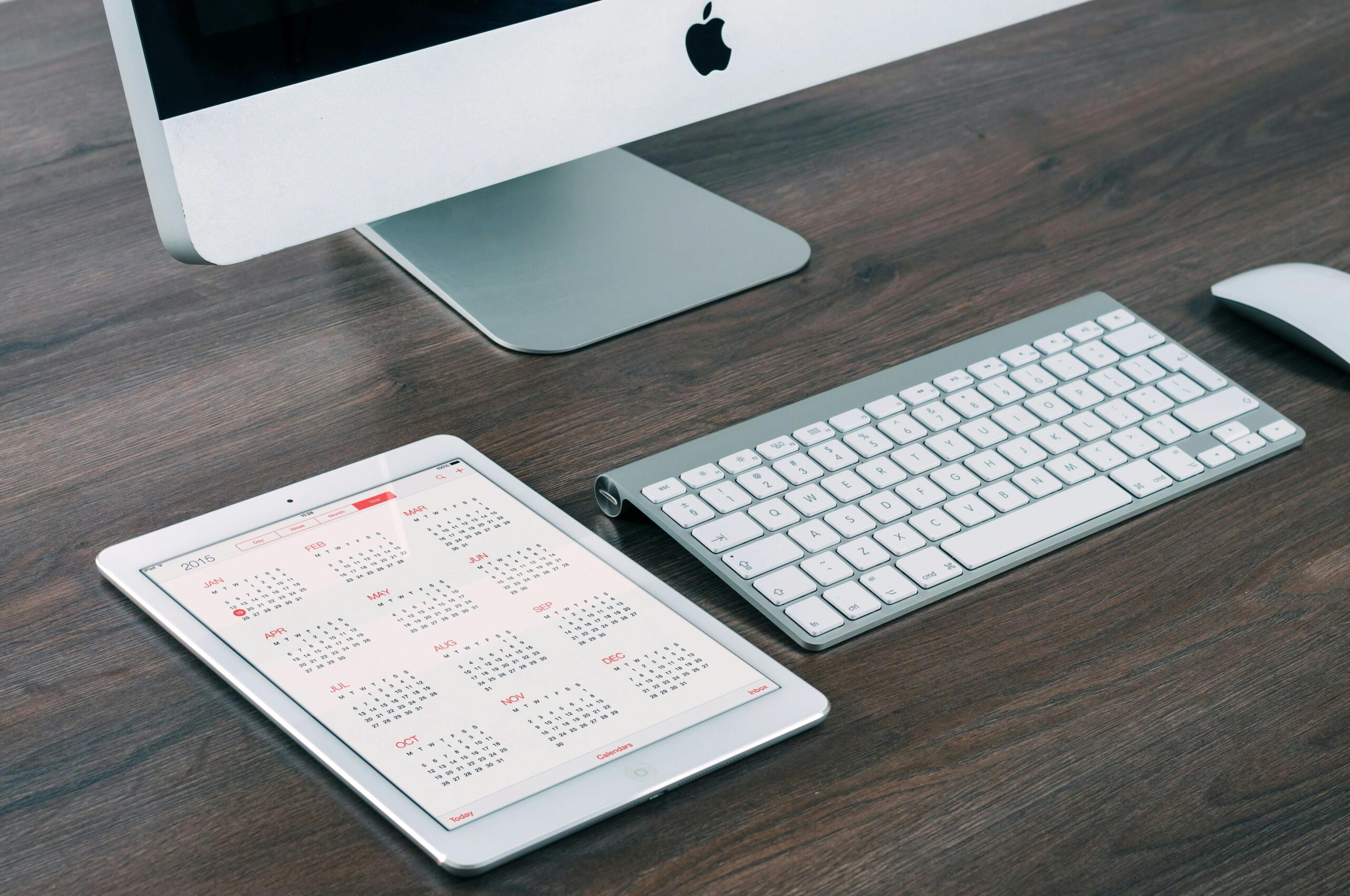 A modern workspace with an Apple computer monitor, keyboard, and tablet displaying a calendar for July 2023.