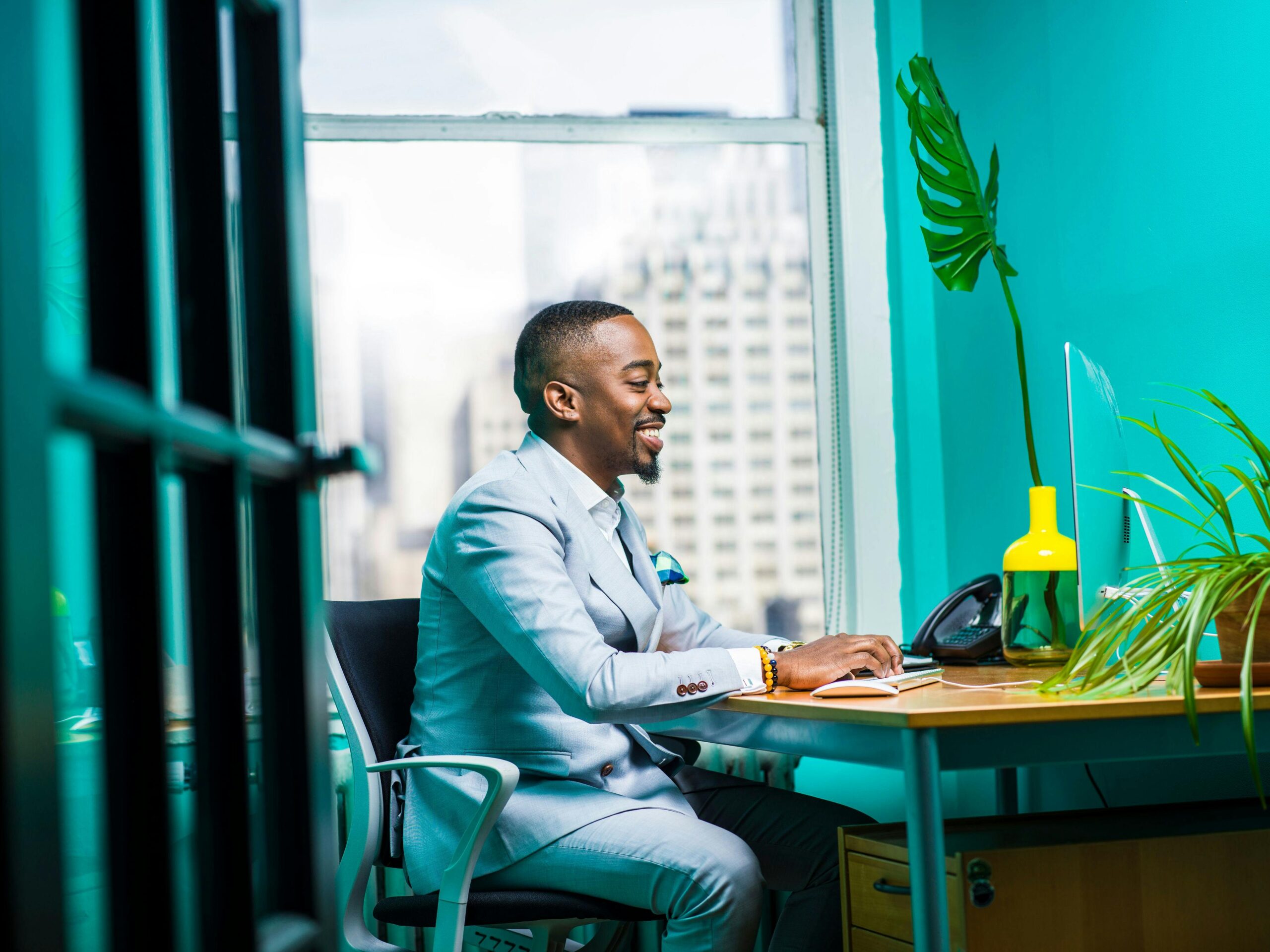 Smiling professional working on a laptop in a bright, modern office setting with green plants and city view.