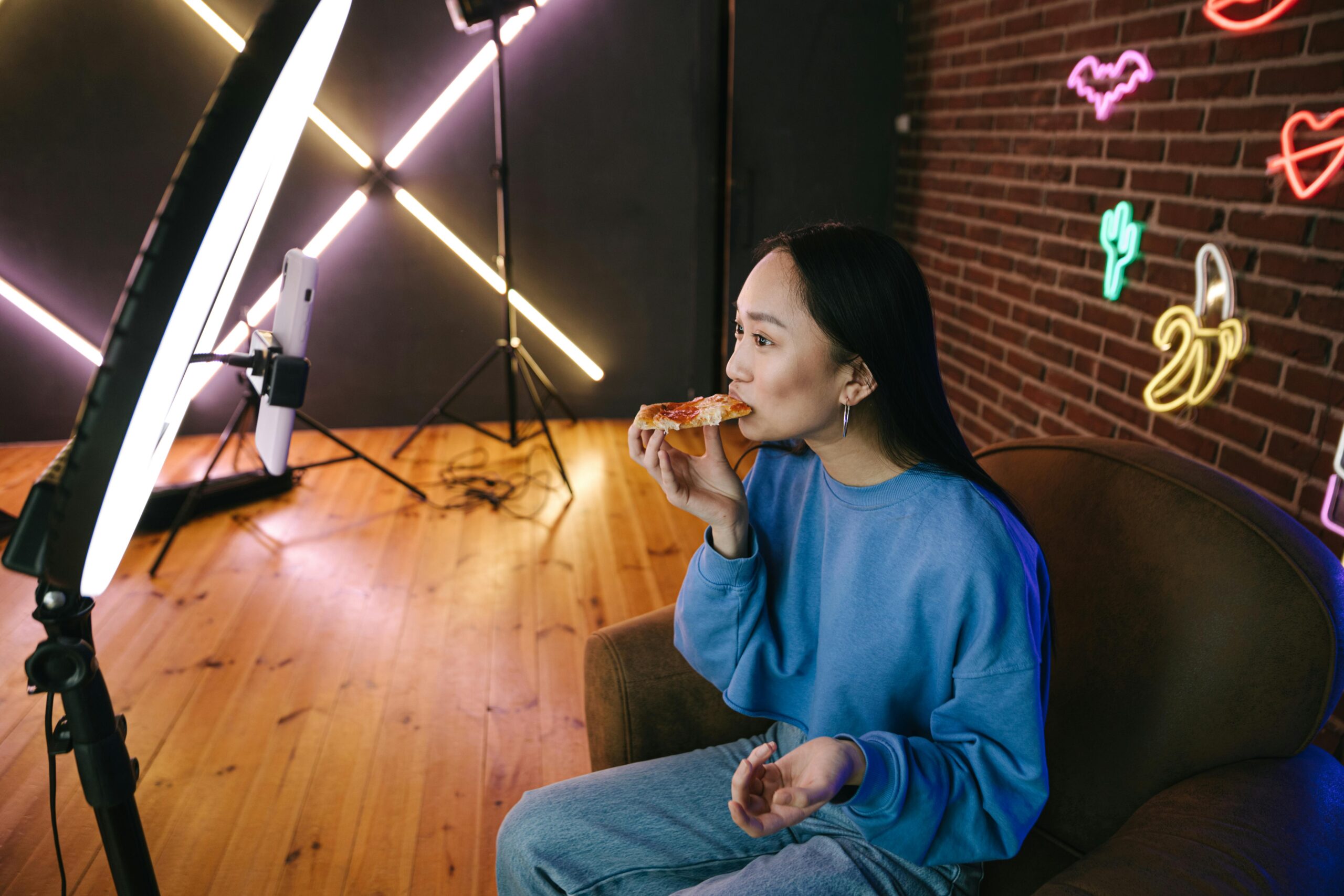 Young woman enjoying a slice of pizza while filming a video with professional lighting setup in the background.