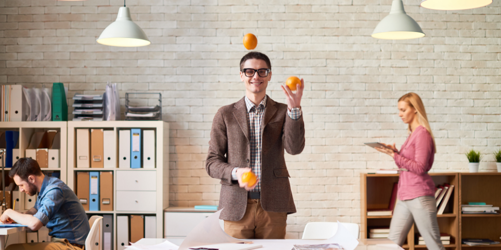 A man in a casual office setting juggling oranges, with colleagues working in the background, indicating a fun and creative work atmosphere.