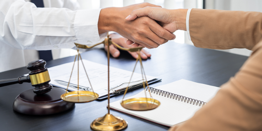 Two people shaking hands over a desk, with a gavel and a balance scale, suggesting a legal or formal agreement.