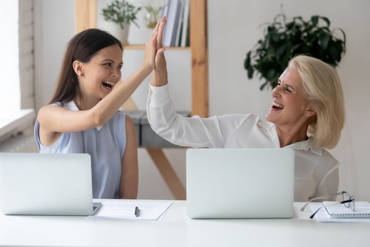 Two women at a desk, one standing, celebrating with a high-five in an office environment.