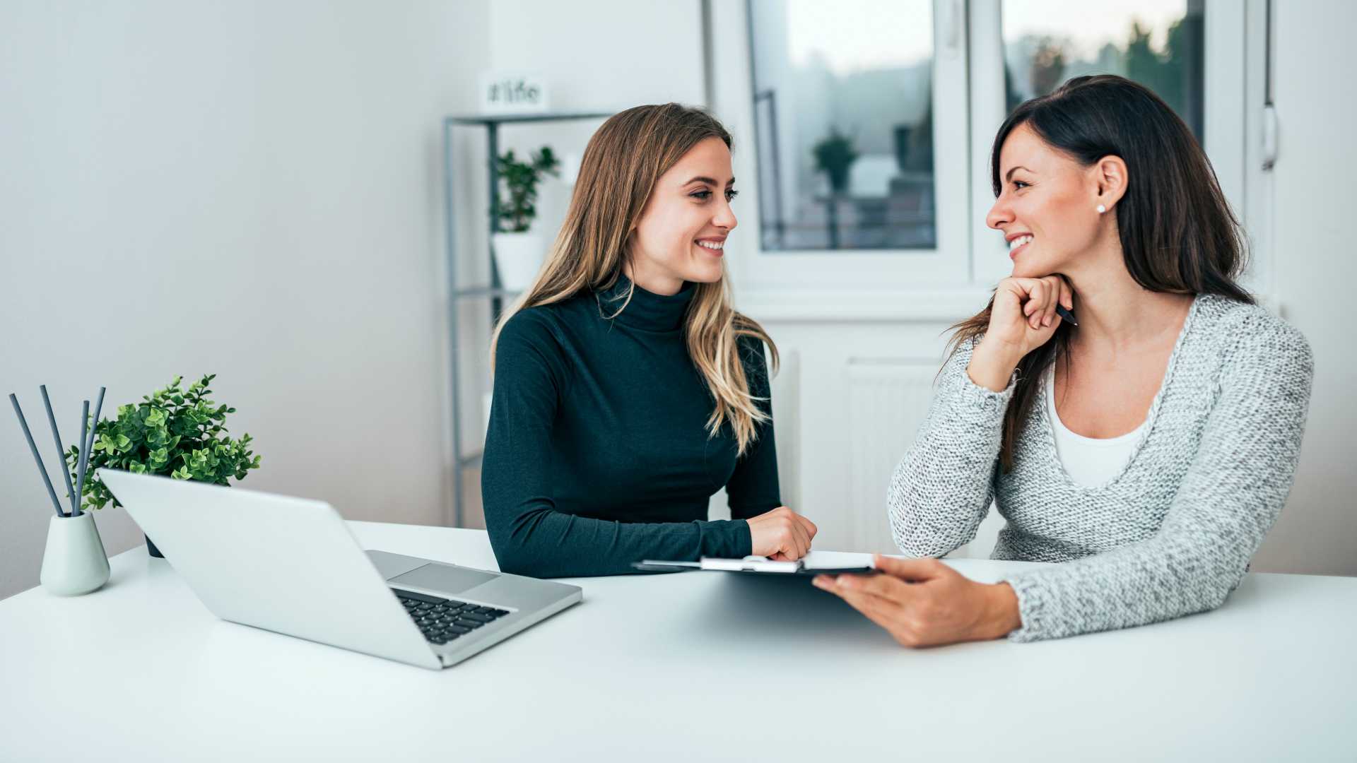 Two women sitting across from each other at a desk, engaging in a conversation with laptops in front of them.