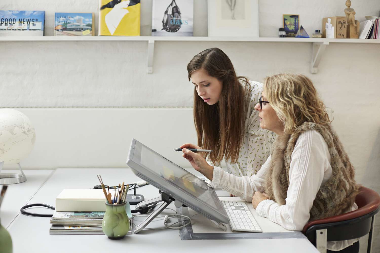 Two women, one younger and one older, are collaborating at a computer workstation with a stylus and digital drawing pad.