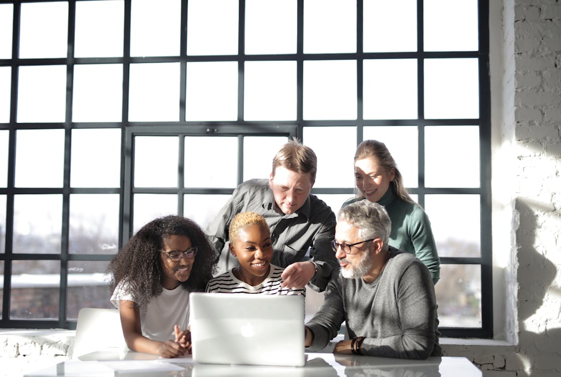 A diverse team of professionals is huddled around a laptop, engaged in a collaborative task in a sunlit room with large windows.
