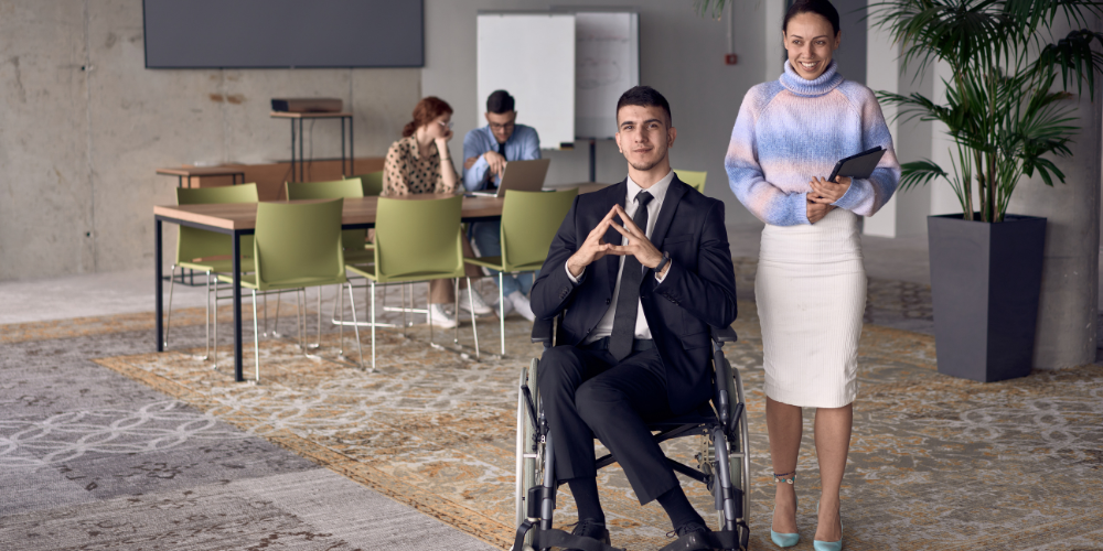 A professional woman stands beside a seated man in a wheelchair in a modern office environment, symbolizing inclusivity in the workplace.