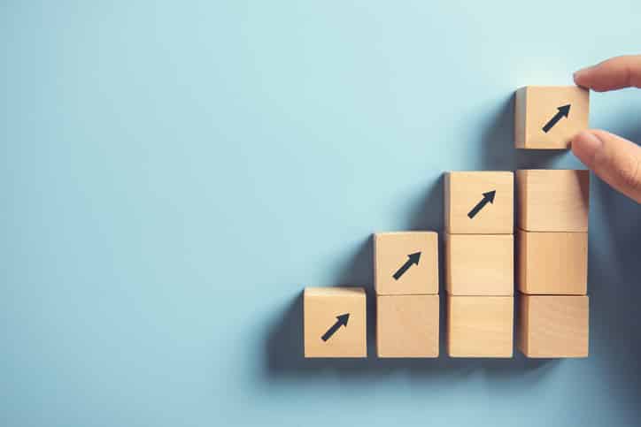 A hand is placing the last of several wooden blocks with arrows pointing upwards, representing growth or progress in a business context.