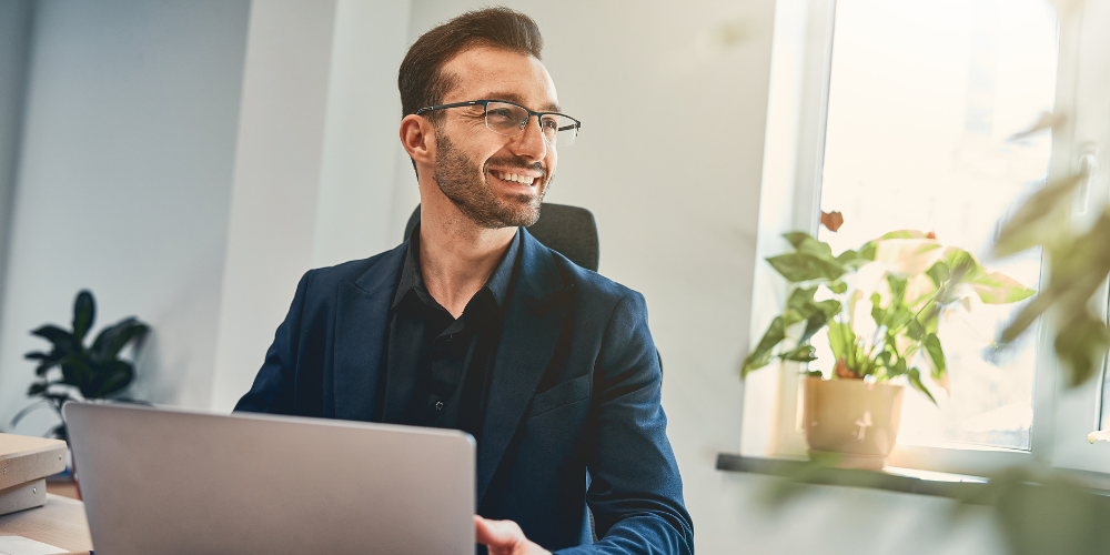 The image shows a professional and cheerful man wearing glasses and a dark suit, sitting at his workspace in front of a laptop, possibly engaging in a video call or work task. A potted plant is visible in the background, suggesting a comfortable office environment with natural elements. The man appears to be smiling, indicating a positive and engaged attitude towards his work.