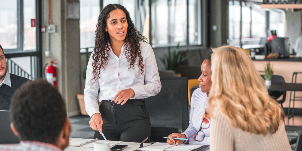A confident woman with long curly hair, standing and speaking at a meeting, with attentive colleagues around a table in an office with large windows.