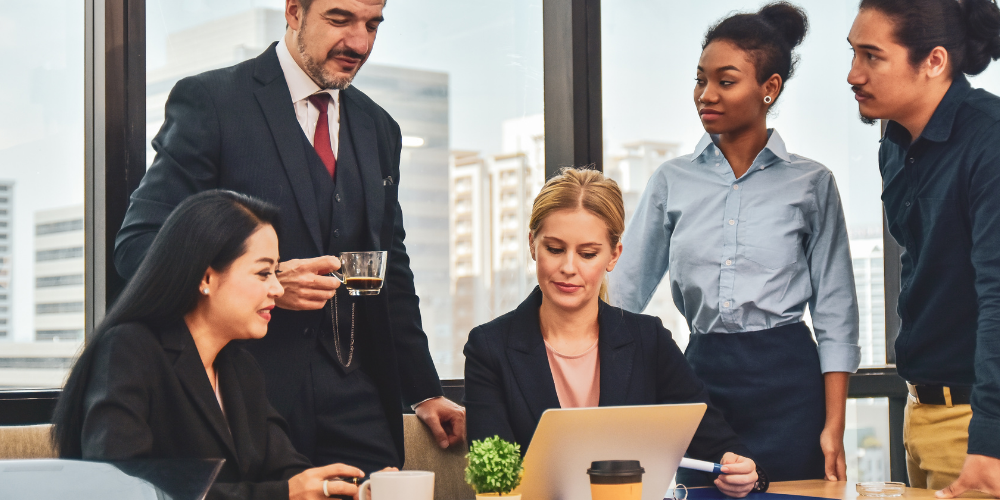 This image shows a diverse group of five professionals around a laptop in what appears to be an office meeting. A man in a suit is standing on the left, holding a cup, next to a seated woman who is smiling and looking at a laptop screen. Another woman is standing in the center, behind the laptop, observing the screen, with two men on the right side—one standing and the other sitting—both looking at the laptop as well. The environment suggests a collaborative and possibly informal business meeting or discussion. The office has large windows, indicating a high-rise building, and the attire of the individuals ranges from business formal to smart casual.
