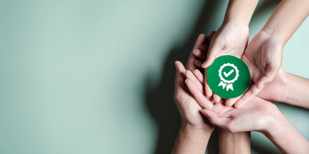 Multiple hands holding up a green badge with a white check mark, symbolizing achievement, quality, or certification.