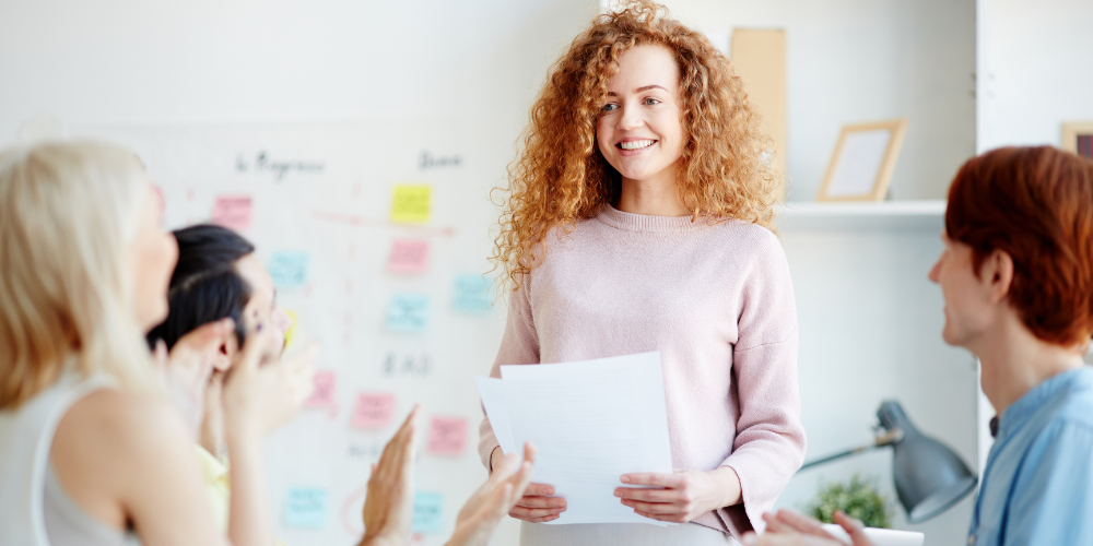 A business meeting with a curly-haired woman presenting to colleagues in front of a whiteboard covered with sticky notes.