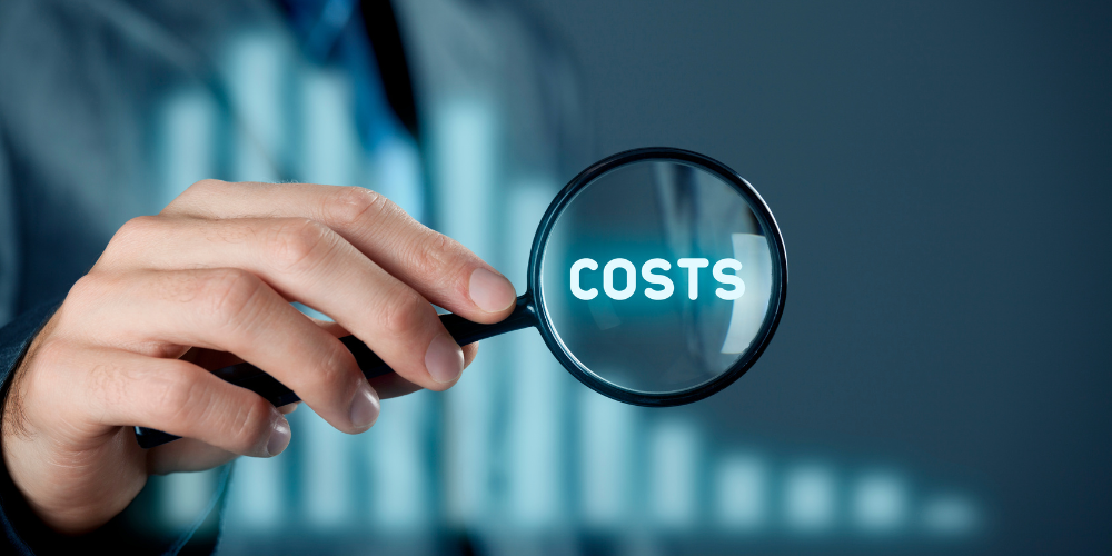 A person's hand holding a magnifying glass over the word COSTS, with a blurred bar graph in the background.