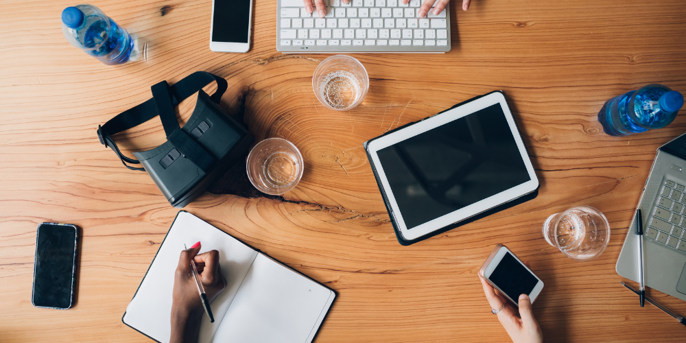 An overhead shot of a workplace table with multiple devices such as smartphones, tablets, a laptop, and hands writing in a notebook, symbolizing a collaborative workspace.