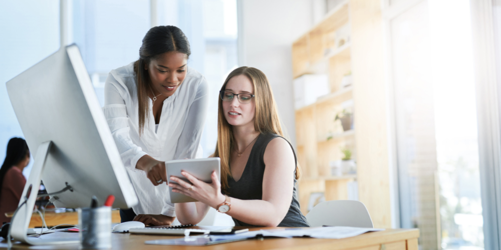 Two professional women, one standing and pointing at a tablet screen, the other sitting at a desk with a computer, collaborating in an office environment.