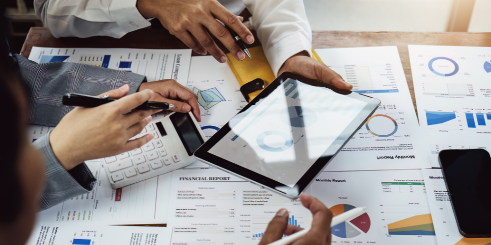 A close-up of a business meeting with hands holding tablets and pens over financial charts and reports, symbolizing team analysis or strategy planning.