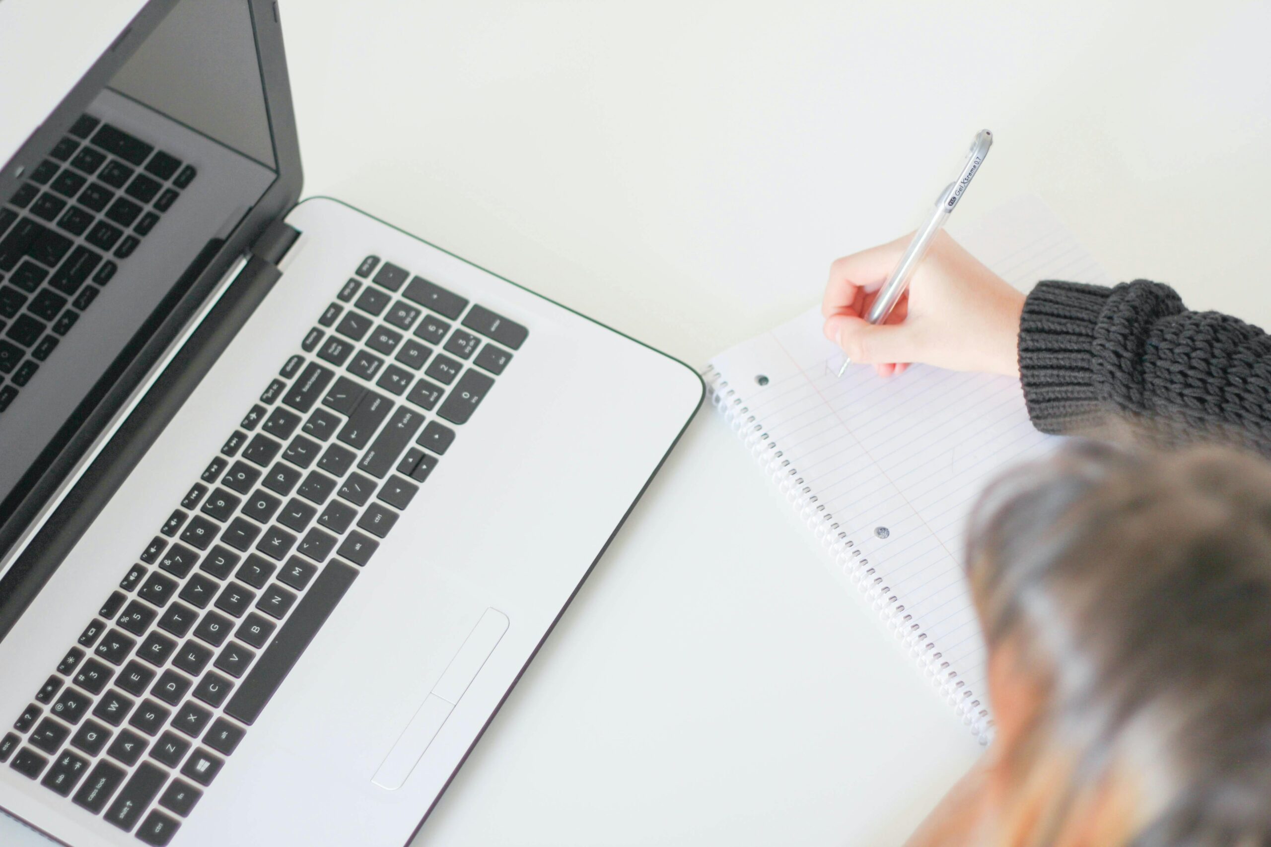 Person writing notes next to an open laptop on a white desk.