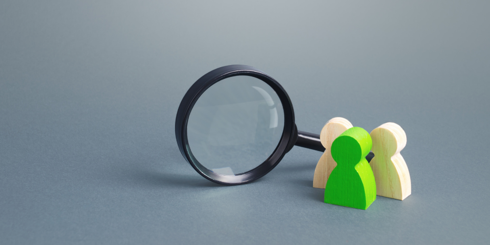 Two wooden figures, one green, standing under a magnifying glass, representing scrutiny, evaluation, or focus on individuals or a group.