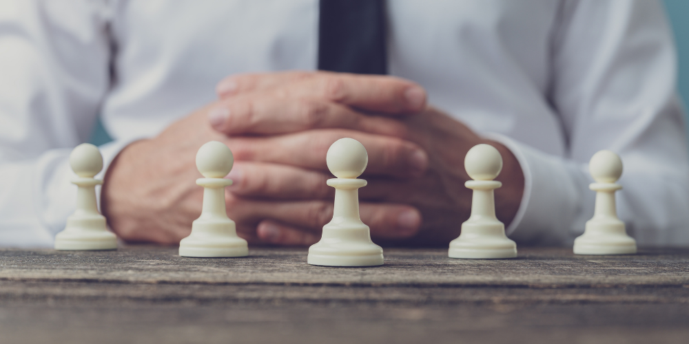 A man in a business suit with folded hands sitting behind a row of chess pawns, suggesting strategy, leadership, or management.