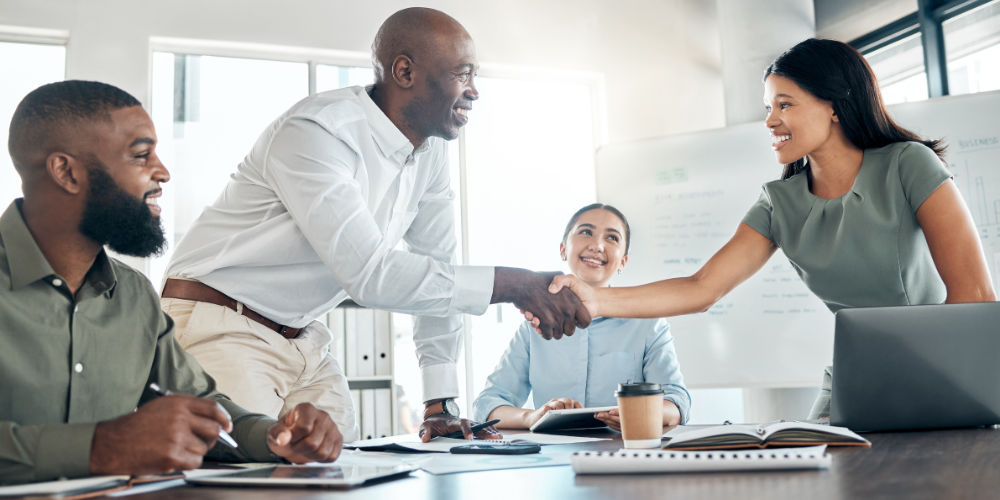 A business team meeting where a man is shaking hands with a woman, signifying agreement or collaboration, with colleagues around them.