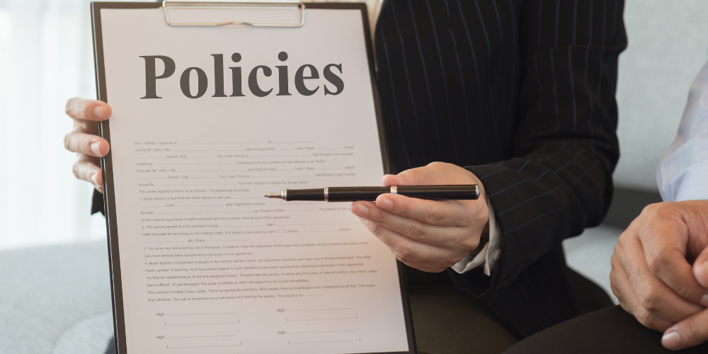 A person holding a clipboard with the title "Policies" and a pen, indicating a review or discussion of company policies or documents.