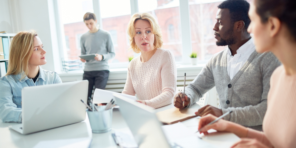 A business meeting in a light-filled office with a diverse team and one person seemingly explaining a point or leading a discussion.
