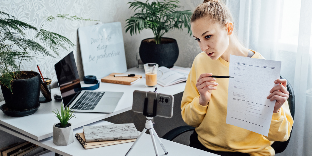 A young woman working from home, reviewing a document while recording or live-streaming, indicating a remote work environment or online content creation.
