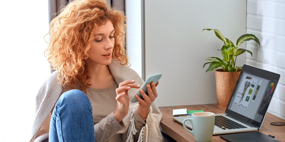 A woman sitting in a cozy indoor setting using a smartphone with a laptop open on the table next to her.