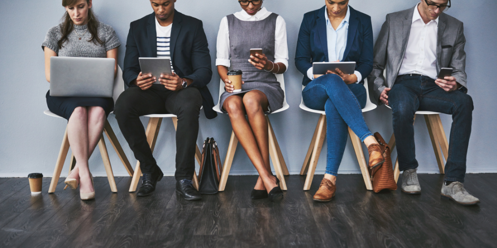 A row of individuals sitting on chairs using various devices such as laptops, tablets, and smartphones, indicative of a waiting area or a tech-engaged group.