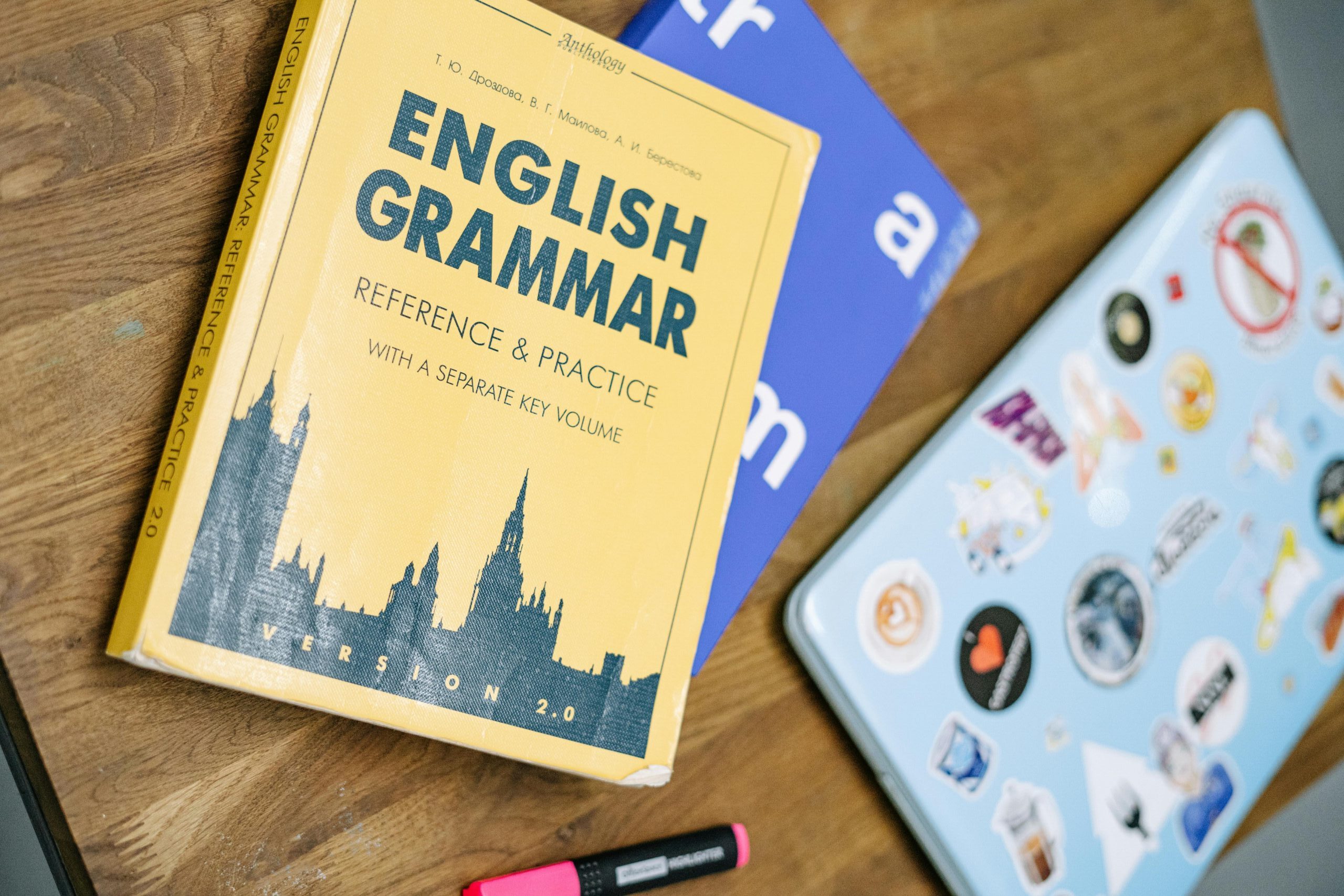 An open English grammar book beside a laptop covered in vibrant stickers, emphasizing the importance of grammar in digital communication.