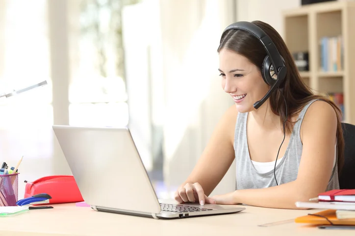 A woman with a headset, smiling as she looks at her laptop screen, which is on a desk with various items like pens and a notepad.