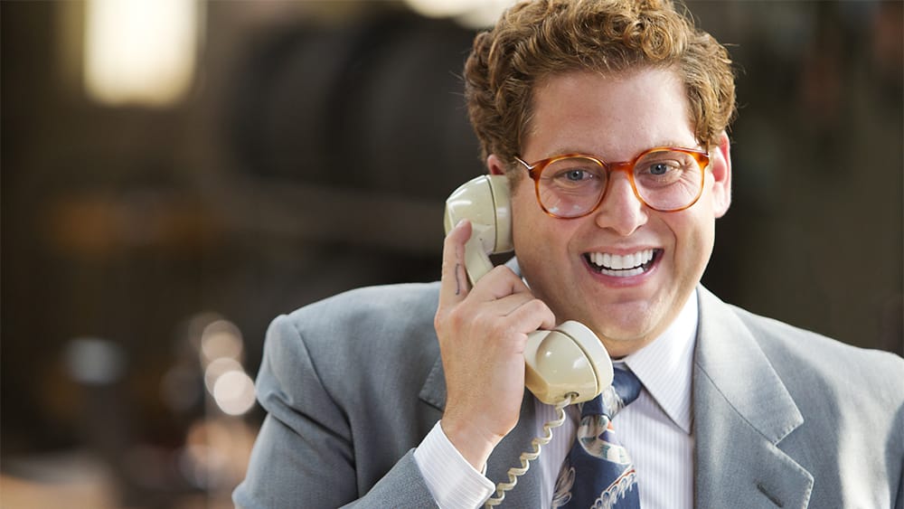 A smiling man with curly hair, wearing glasses, and a business suit is holding a cream-colored rotary phone to his ear.