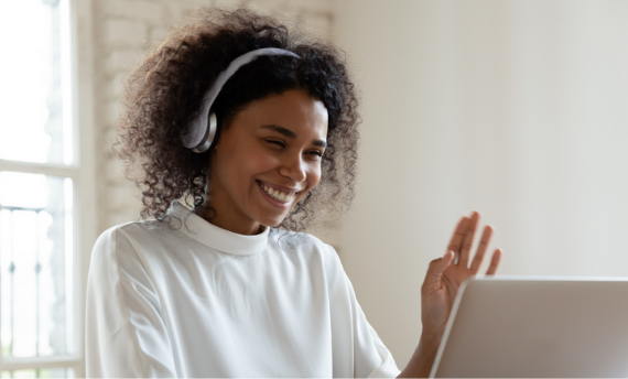 A cheerful woman wearing a white headset, waving at her laptop screen during what appears to be a video call.