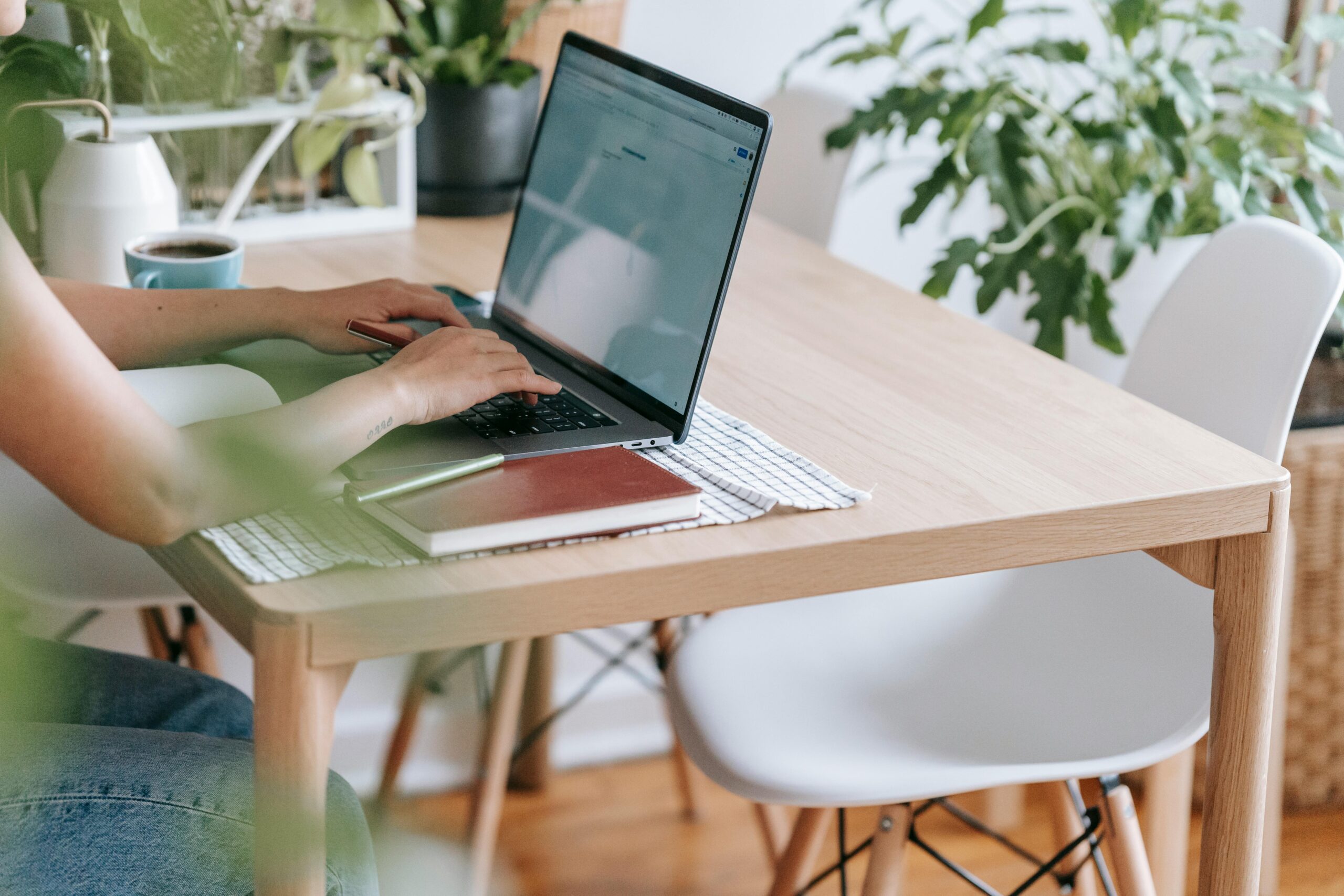 A person typing on a laptop in a bright home office space with green plants and a white desk.