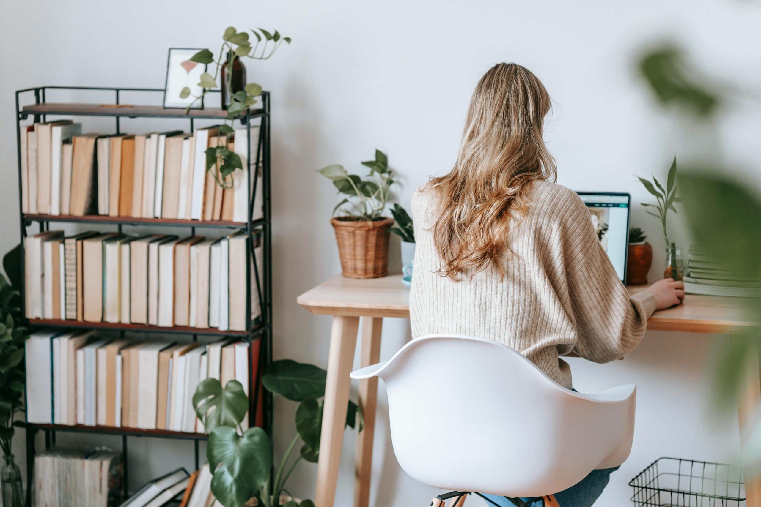 A person with long hair working on a laptop at a home office desk surrounded by plants and a bookshelf. She might be wondering what are the rates for virtual assistants.
