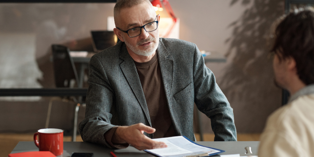 An older man with glasses in a plaid blazer discussing over a table with a younger person, not visible in the image.
