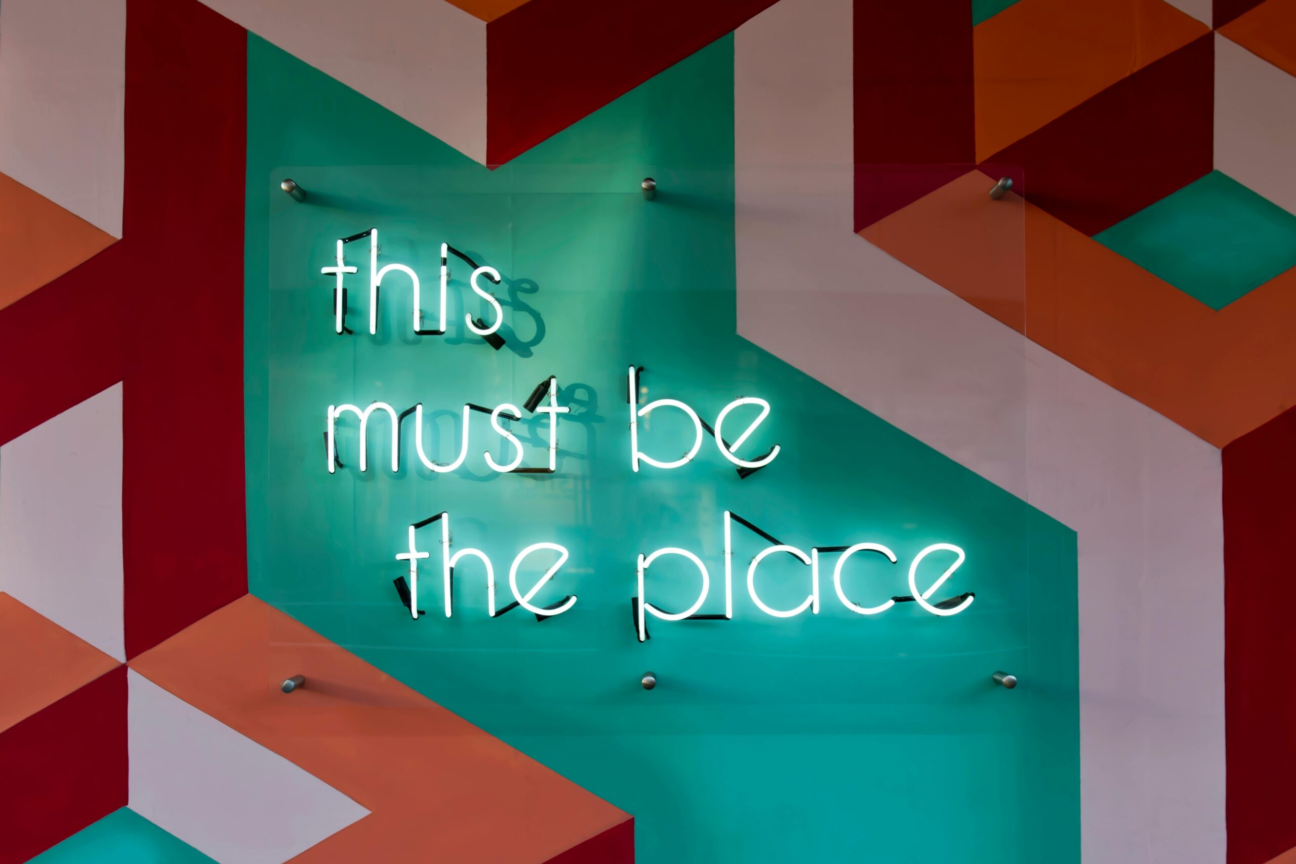 Neon sign reading "this must be the place" against a geometric background, resonating with the concept of what is recruitment outsourcing.
