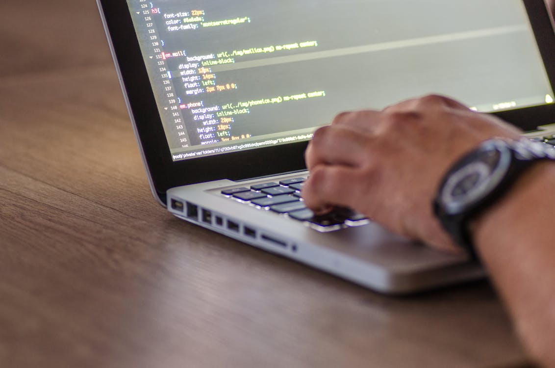 A close-up of a person's hand typing on a laptop keyboard with code visible on the screen.