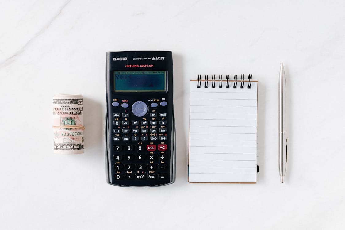 A Casio scientific calculator, a roll of US dollar bills, and a notepad with a pen on a white surface.