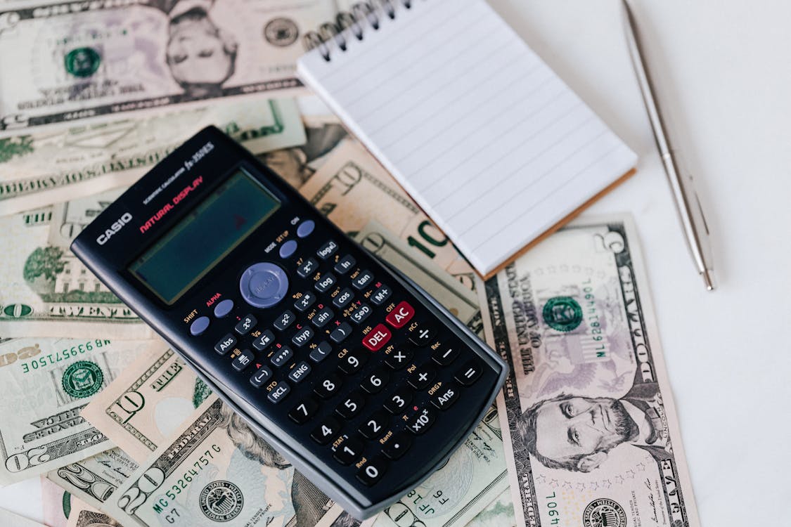 A Casio calculator on top of a spread of US dollar bills next to a notepad and pen.