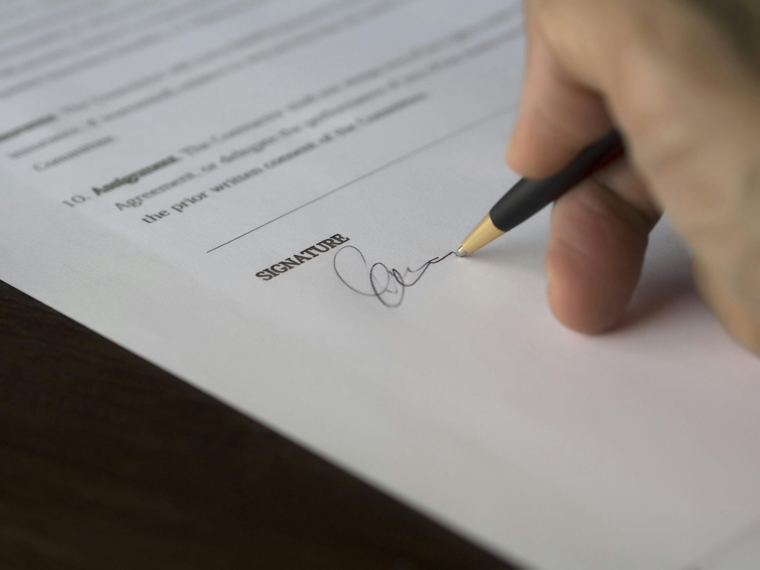 A hand signing a document, possibly a contract for independent contractor sample, with a black pen on a wooden surface.