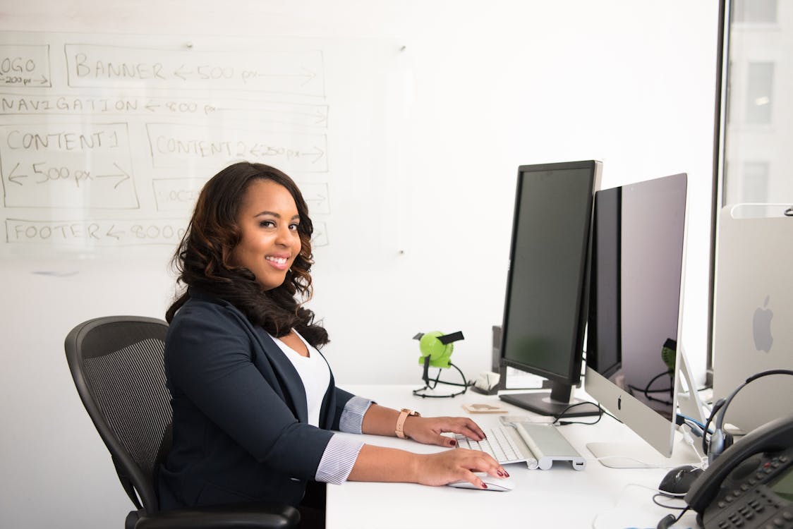 Confident executive assistant working at her desk with dual monitors and a whiteboard with website design layout in the background.