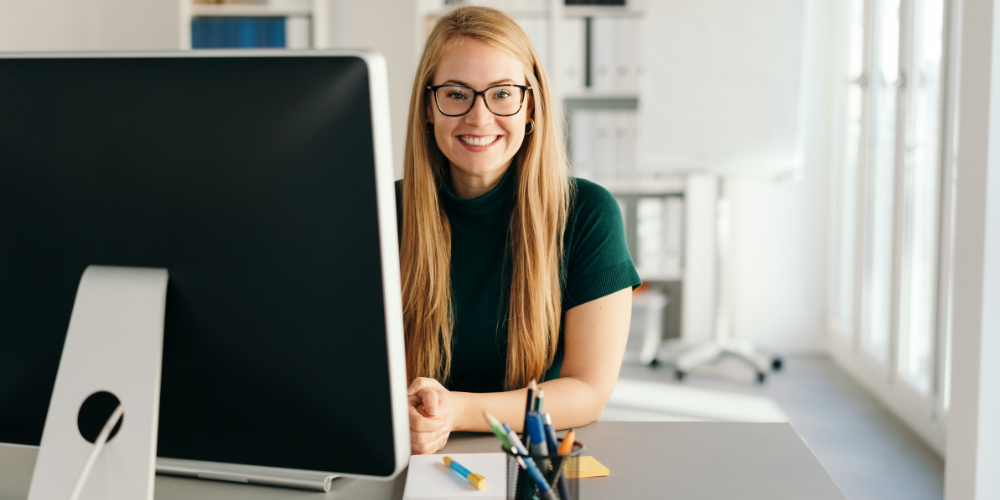A happy woman with glasses sitting at her desk in front of a computer.