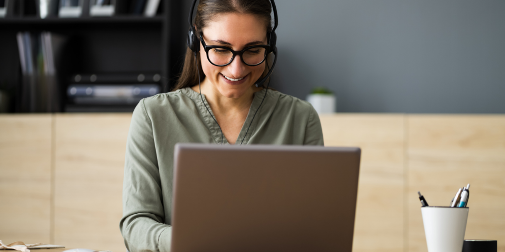 A virtual assistant with headphones smiling while looking at her laptop.